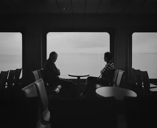 A black and white scene inside a ferry or ship lounge, where two men sit facing each other at a small table, silhouetted against large windows overlooking the water.