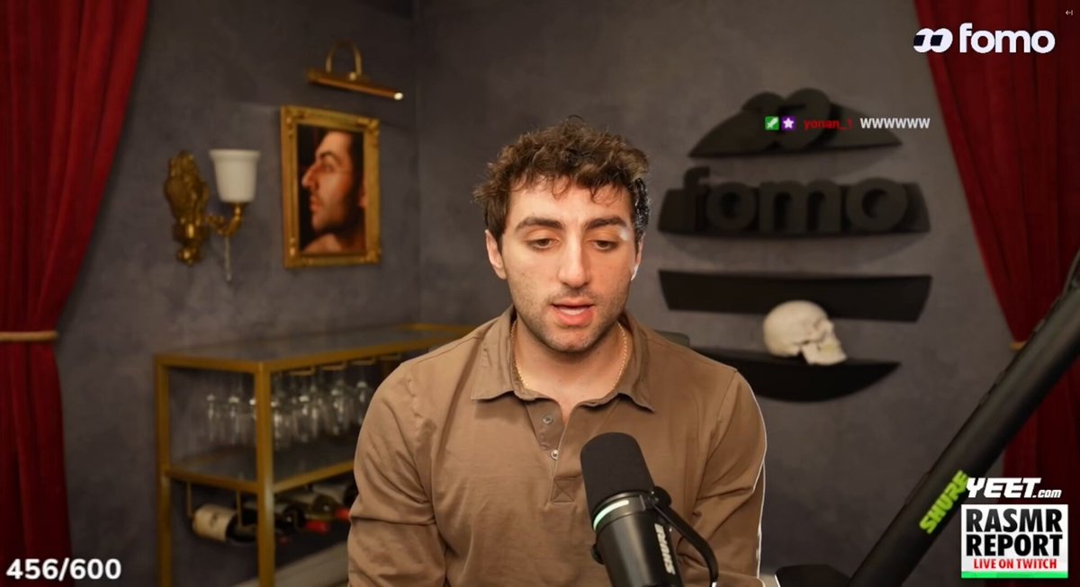 The image shows a man wearing a brown shirt sitting behind a microphone at a podcast studio. The host of the show appears to be asking him questions, possibly about his thoughts or opinions on a topic related to finance or technology. The background features a red curtain with a painting hanging above it and a wine rack next to it.
