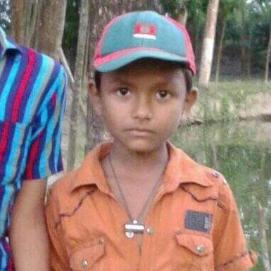 A photo of a young boy wearing an orange shirt and a teal and red cap with the Bangladesh flag, standing outdoors with another person partially visible beside him.