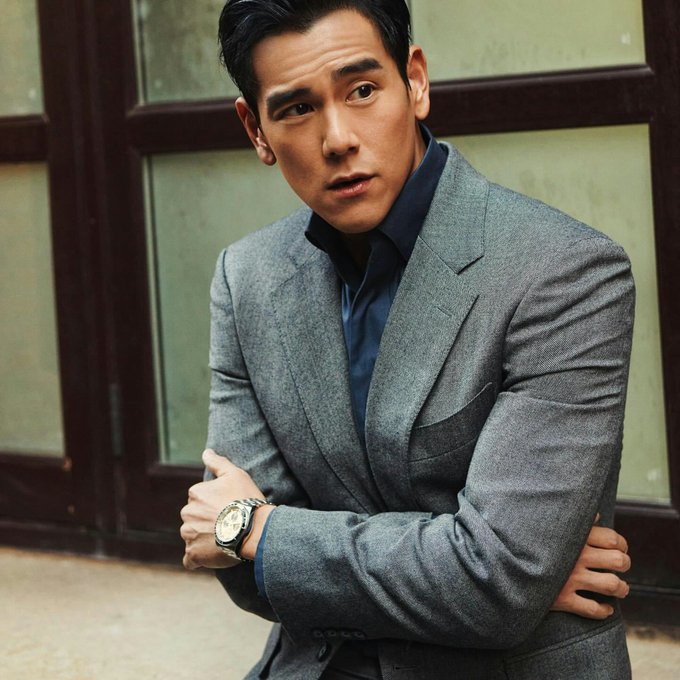 Editorial portrait of a man in a grey suit and blue shirt wearing a silver watch, posed with crossed arms against a paneled background.