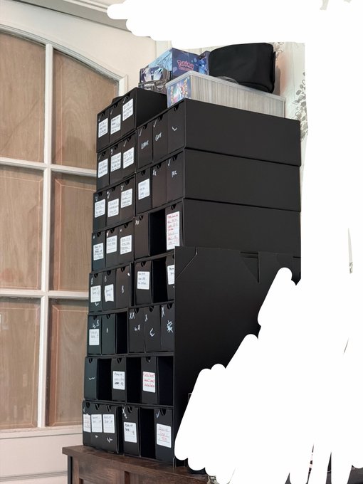 A tall stack of black storage boxes with white labels sitting in front of wooden cabinets.