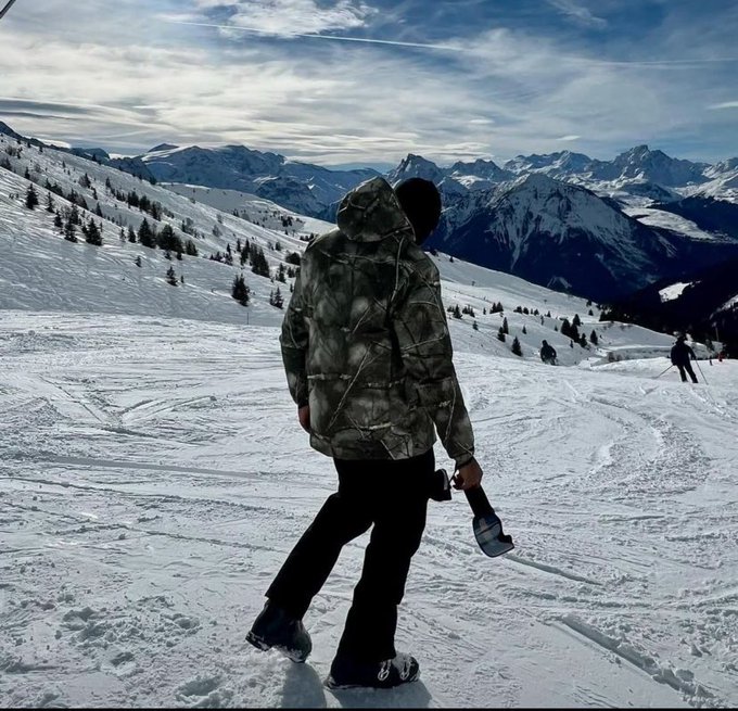 A person wearing a camouflage hooded jacket and dark pants walks through a snowy mountain landscape carrying a snowboard, with distant peaks visible under a partly cloudy sky.