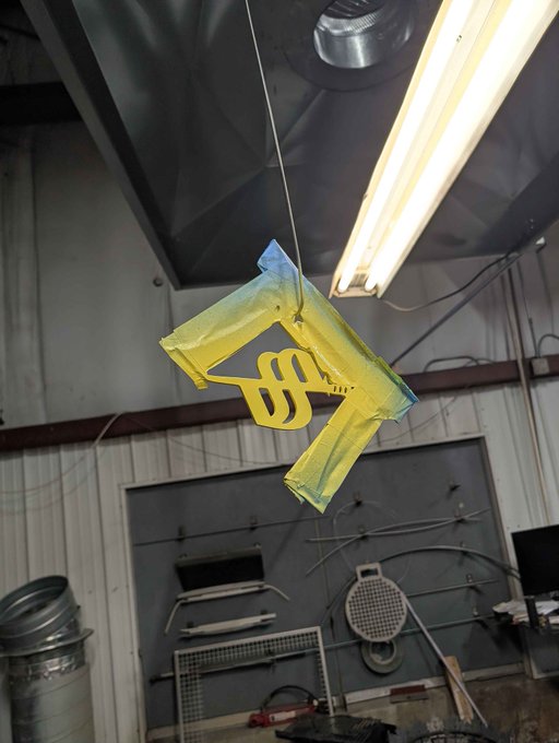 A yellow toy gun hangs from a string in what appears to be a workshop or garage, illuminated by a fluorescent light overhead. The whimsical, makeshift setting complements the playful "Gm from the Dojo" greeting, evoking a fun, DIY atmosphere fitting for a casual morning send-off.