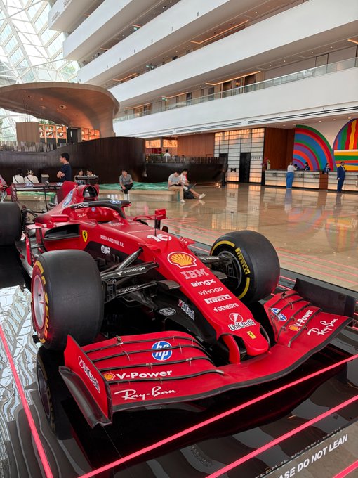A red Ferrari Formula 1 race car with sponsor logos including Shell, IBM, and Ray-Ban is on display inside a modern multi-level building lobby.