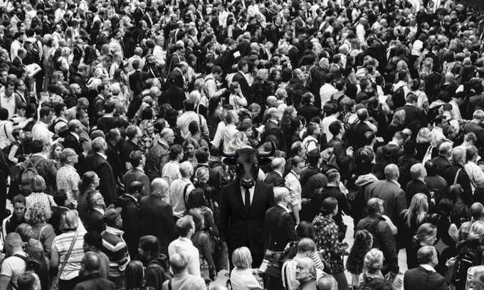 A black and white photograph showing a dense crowd of people, with a central figure wearing a black suit, white shirt, and a dark animal (possibly a panther) mask.