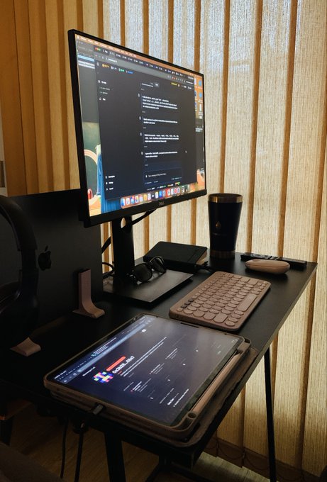 A modern desk setup with a large monitor, an iPad, keyboard, mouse, tumbler, and a Mac Studio against vertical wooden blinds.