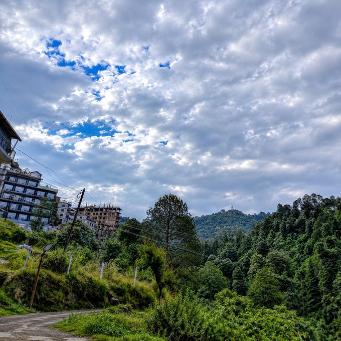 Scenic landscape photograph showing a cloudy sky over a hillside town with green forested mountains and buildings on the left.