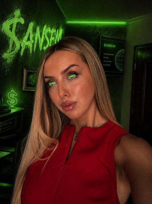 A selfie of a blonde woman in a red top in a green-lit room with 'SANSEM' written on the wall, a dollar sign lamp, and a framed quote about the movement being real and the future being a system.