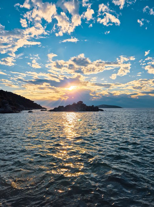A scenic sunset over the ocean, with sun rays breaking through scattered clouds and reflecting on the rippling water, with dark rocky islands visible in the distance.