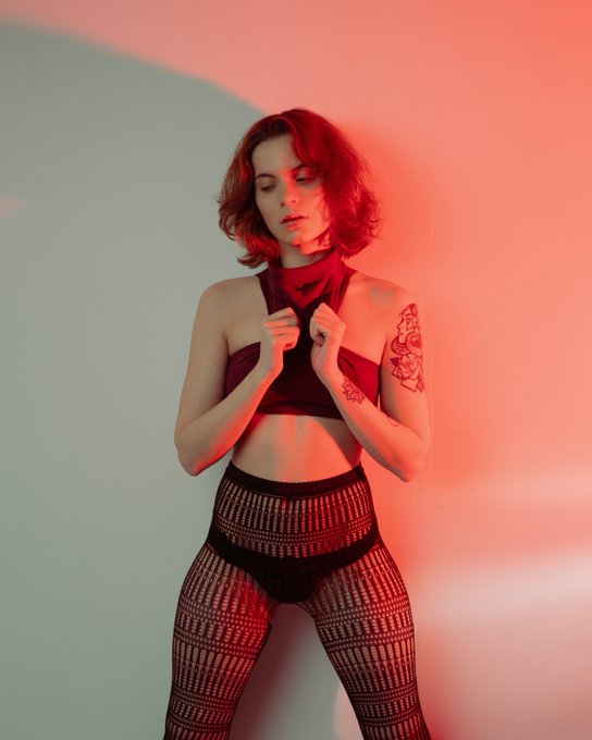 A woman with short reddish hair poses in red and black lingerie against a wall bathed in red light, with a floral tattoo visible on her left arm.