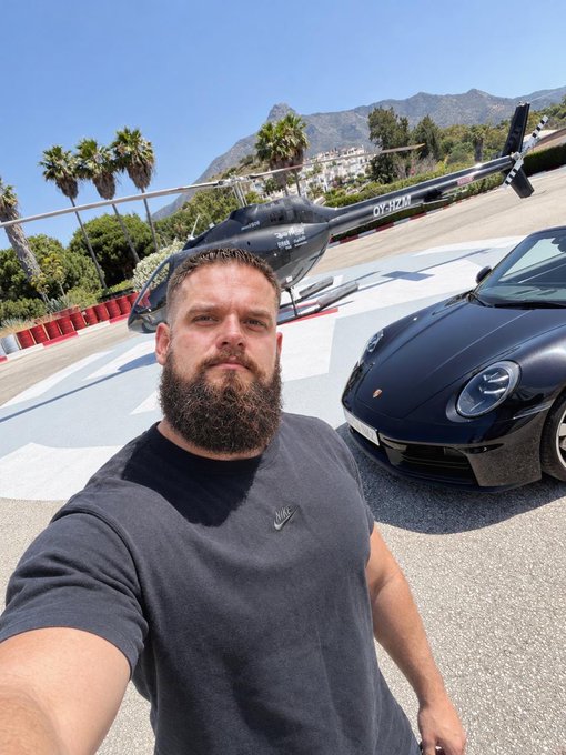 A bearded man takes a selfie in front of a black Porsche and a helicopter marked "OY-HFH," with palm trees and mountains in the background—visually illustrating the "GM, my friends" (Geneva Motor Show / luxury lifestyle) vibe.