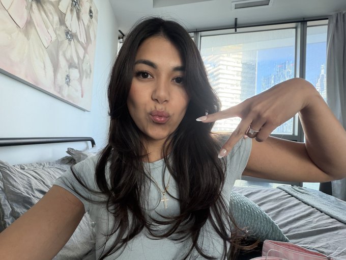 A young woman with long dark hair taking a selfie in a bedroom, wearing a gray t-shirt and cross necklace, posing with puckered lips and holding her hand up near her face.