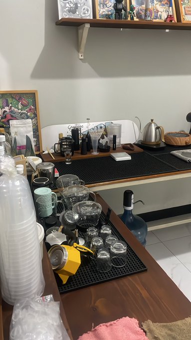 A home bar/counter setup featuring various glassware, a yellow moka pot, bottles of liquor, a kettle, and decorative items including framed art and a Gandalf figurine on a shelf above.