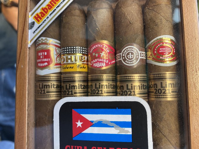 A wooden box containing Cuban cigars from brands like Hoyo de Monterrey, H. Upmann, Partagas, Montecristo, and Romeo y Julieta, marked as 'Edición Limitada 2021' with a Cuban flag sticker.