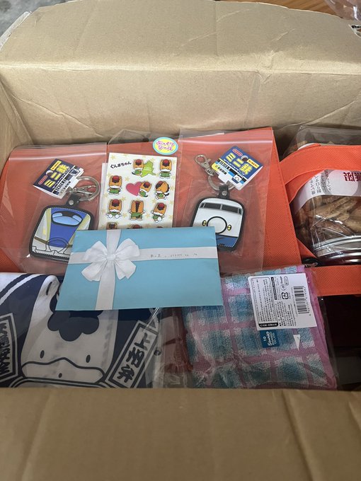 An open cardboard box filled with various items in plastic packaging, including an orange pouch, a light blue card with a white bow, keychains, and a pink patterned cloth.