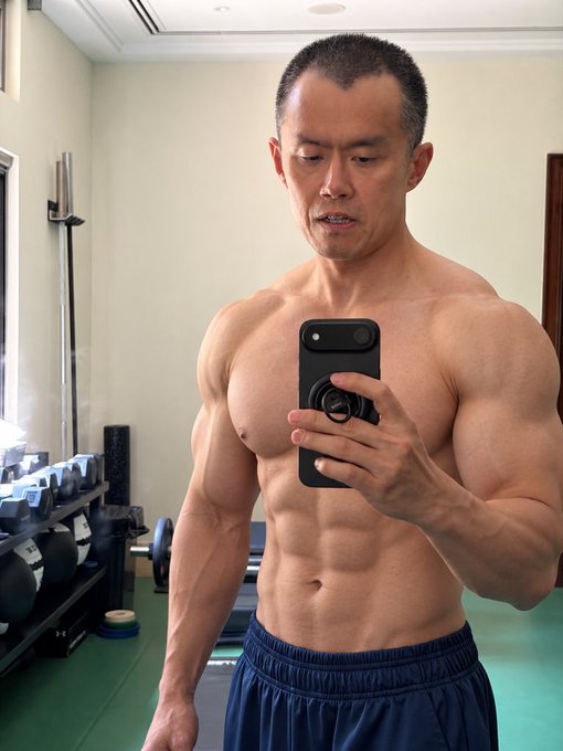 A shirtless, muscular man with a buzz cut takes a mirror selfie in a gym while holding a black smartphone, wearing navy blue athletic shorts.