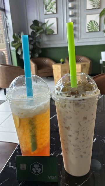 Two iced bubble teas—one a vibrant orange jasmine tea with a blue straw and the other a creamy matcha milk tea with boba and a green straw—sit on a black table next to a green Crypto.com Jade Pro Visa card, matching the post about enjoying a refreshing drink and earning $CRO cash back at a Vietnamese restaurant.