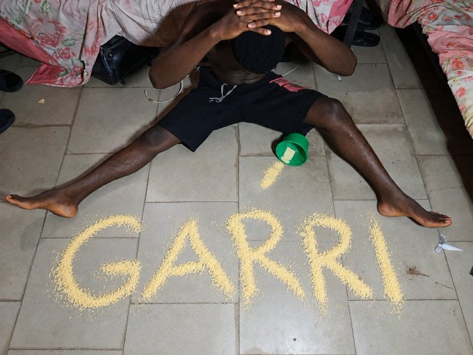 A shirtless man sits on a tiled floor with his hands clasped over his bowed head in a pose of devotion, as the word "GARRI" is spelled out in granular powder in front of him, with a tipped green cup nearby. The image visually reinforces the post's playful devotion to the $GARRI memecoin, with the man humorously using his last bit of the staple West African food as a tribute to the token.