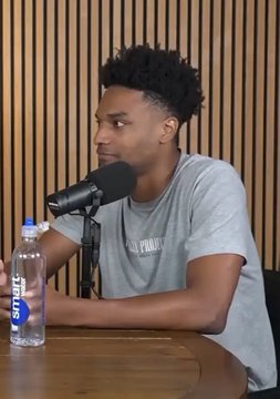 A young man sits at a wooden table speaking into a microphone during what appears to be a podcast recording, with a water bottle labeled "smartwater" beside him and a slatted wooden wall in the background. The image likely shows the speaker referenced in the post, Ansem, sharing his bullish outlook on crypto's mainstream adoption by 2028 through mobile social trading apps.