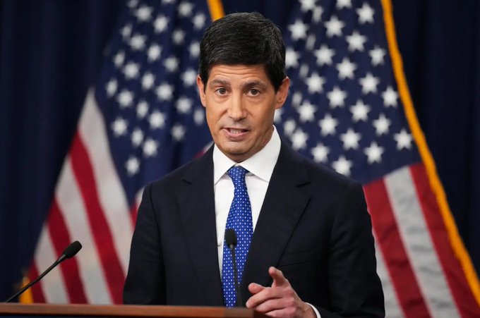 A man in a dark suit and blue patterned tie speaks at a podium with American flags in the background, appearing to be at a political or government press event.