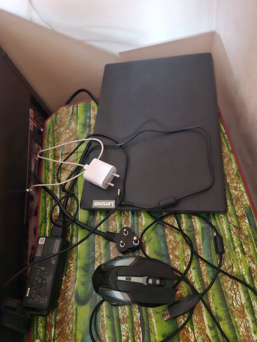 A black Lenovo laptop sits closed on a green-patterned surface, accompanied by a white power adapter, a gaming mouse with cables, and a power brick.