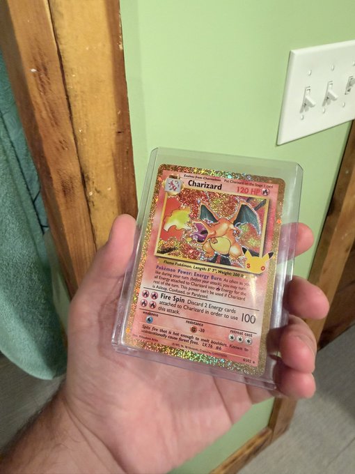 A hand holding a holographic/rainbow Charizard Pokémon card encased in a protective sleeve, displaying its Fire Spin attack (100 damage), 120 HP, and first edition stamp.