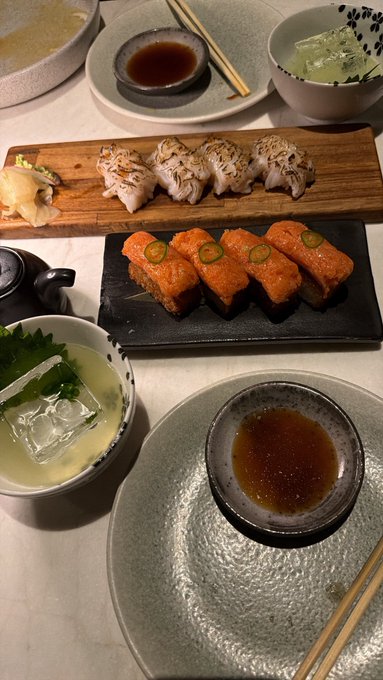 The image shows a dining table with various sushi dishes, including plates of fish and other food items such as seaweed salad and dipping sauces. The table also has chopsticks placed next to some bowls containing additional food. A bowl filled with ice is present on one side of the table. The scene appears to be set for a sushi night or gathering where guests can enjoy different types of sushi along with accompanying dips, creating an interactive dining experience.
