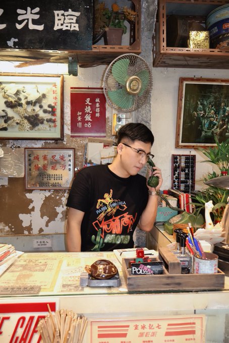 A young man wearing glasses and a black graphic t-shirt holds a green rotary phone to his ear inside a cluttered vintage shop filled with Chinese signage, artwork, and memorabilia. The nostalgic setting with its old-school decor echoes the post's theme of analyzing long-term market trends through moving averages, suggesting a patient, time-tested approach to catching the "bottom" before a breakout.