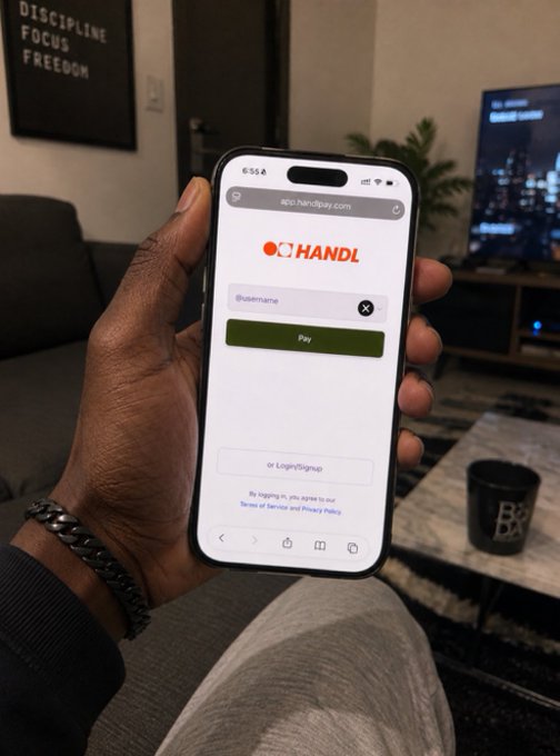 A hand holds a smartphone displaying the HandlPay login page at app.handlpay.com, illustrating the app that allows users to get paid via direct messages. The cozy living room backdrop with "DISCIPLINE FOCUS FREEDOM" wall art adds a relaxed vibe to this fintech moment.