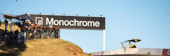 A group of spectators gathers behind a large black "Monochrome" branded sign at what appears to be an outdoor racing or sporting event under a clear blue sky. The image serves as promotional sponsorship for the @monochrome_en giveaway, with the company's branding prominently displayed alongside the $50 USDT prize announcement.