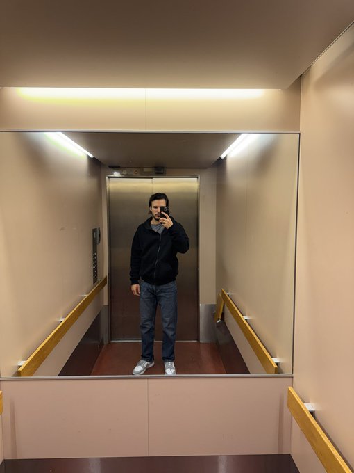 A mirror selfie of a man wearing a black jacket and jeans standing in an elevator with pink walls and yellow handrails.