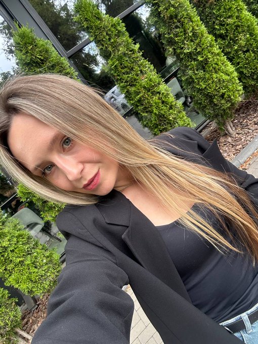 A blonde woman in a black blazer and dark top takes a selfie outdoors, with green shrubs and a building visible in the background.