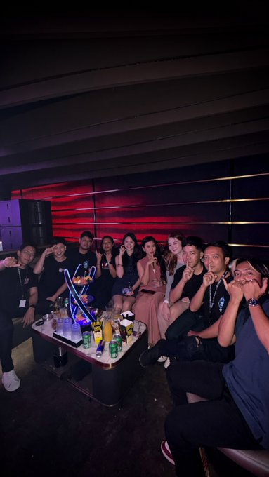 A dimly lit photograph of a group of young adults posing together on a black leather sofa around a low table in a lounge or club setting with red accent lighting.