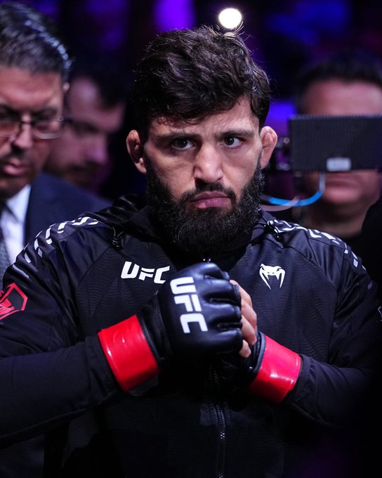 A bearded UFC fighter wearing black Venum gear and UFC-branded gloves stands with his hands clasped together, his intense gaze fixed forward in what appears to be a pre-fight walkout moment. The image aligns with the post promoting Arman "Arman UFC," likely ahead of his bout at UFC 331.