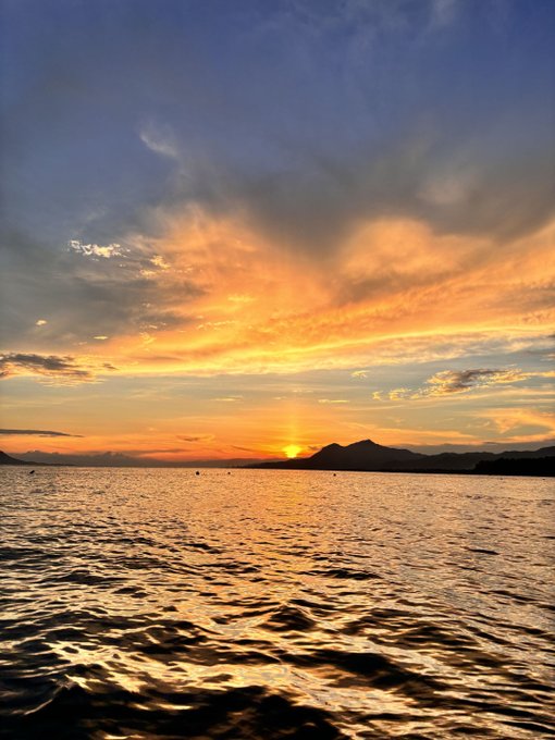 A vibrant sunset with orange and blue clouds reflects across a calm body of water with mountains silhouetted on the horizon.