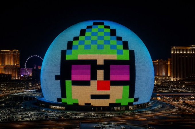 The Las Vegas Sphere is illuminated at night with a massive pixel-art display of a green-haired face wearing pixelated sunglasses, dominating the iconic curved venue. The colorful 8-bit style portrait transforms the futuristic landmark into a giant retro video game character, playfully matching the caption's humor about accidentally creating a memorable landmark.
