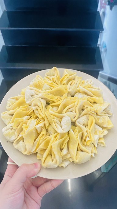 A hand holds a white plate filled with neatly arranged, handmade wonton wrappers (hoành thánh) ready for cooking. The folded yellow dough pieces show the effort put into preparing the wontons, matching the caption about wrapping them at home.