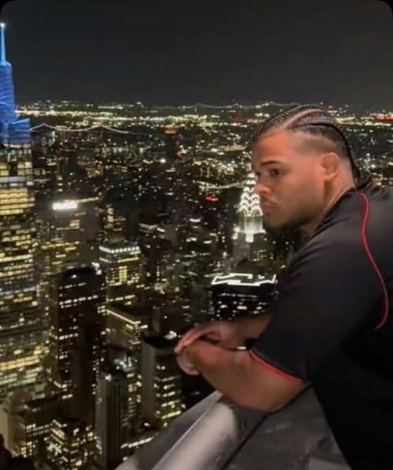The image shows a man with braids standing at the top of a building, looking out over a city skyline at night. The man appears to be posing for a photo or enjoying the view from his vantage point. In the background, there are several tall buildings that create an impressive backdrop for the scene. The post also includes some text about "good morning CT Happy weekend Touch some grASS today and stack more $RIVER points". This message seems unrelated to the main subject of the image but adds context to the overall setting.