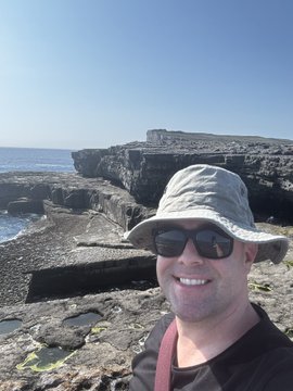 The image features a man wearing sunglasses and a hat standing on a rocky cliff overlooking the ocean, with his face turned towards the camera. The photo has been captioned "Where's the most unique place you've ever bought #SPX690? I just bought at the worm hole on Inishmore, Galway... But I didn't buy wormholed SPX! Always on ETH and withdrawn to Cold Storage LFG".