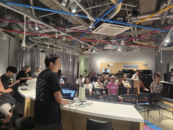 A man stands presenting to a seated audience in a tech-style room with exposed colorful pipes, using a laptop while attendees watch attentively.