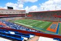 A panoramic view of a large football stadium with green field and orange seating, viewed under a cloudy sky.