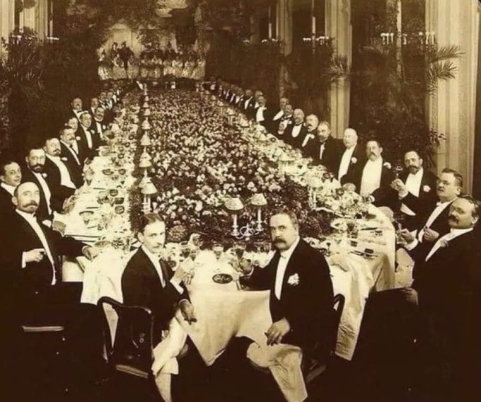 A sepia-toned vintage photograph showing a large formal banquet with dozens of men in tuxedos seated along a long, flower-decorated table in an ornate hall.