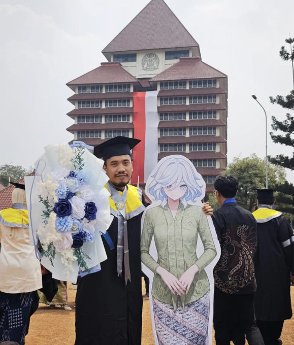 A graduate in cap and gown holding a bouquet of blue and white flowers stands next to a cardboard cutout of an anime-style female character, with a large building featuring a red and white facade in the background.