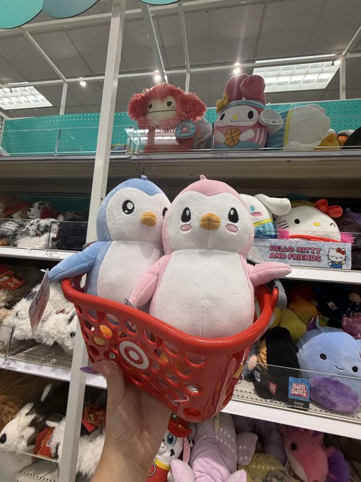A hand holds a red Target shopping basket containing two plush penguins, one blue and one pink, in a Target store aisle stocked with plush toys including Hello Kitty and Friends merchandise. The image captures the moment of finding Pax Pengu and Polly, the Pudy Penguins characters mentioned in the post, at Target.
