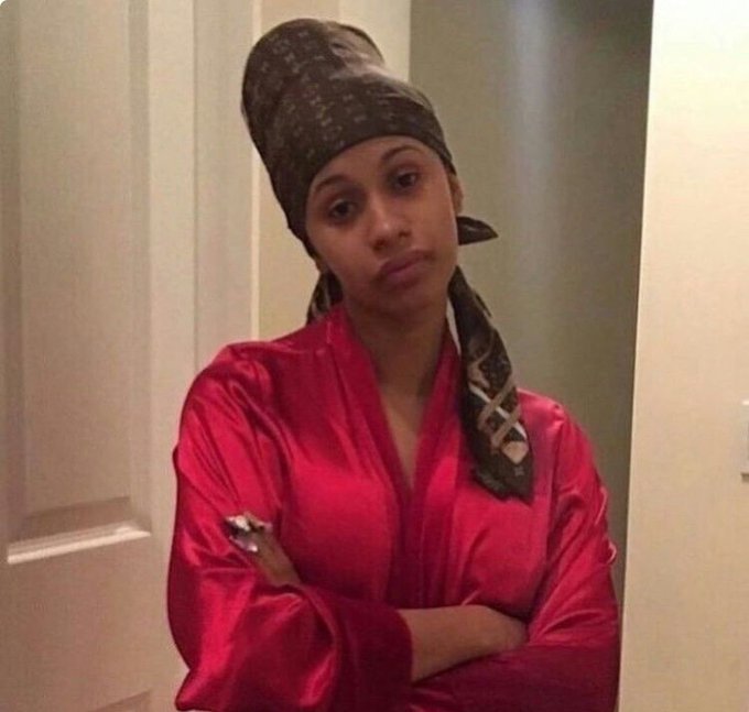 A young woman wearing a red satin robe and a Louis Vuitton patterned headwrap stands with arms crossed in a home setting.