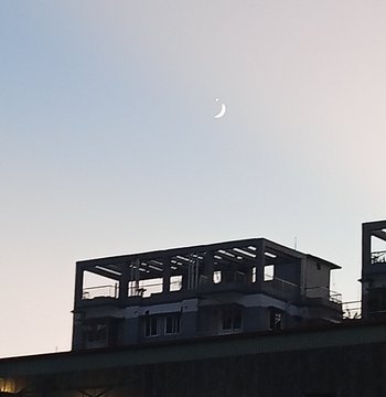 A serene photograph of a crescent moon in a pale sky at dusk or dawn, with the silhouette of a rooftop structure visible in the lower portion of the frame.