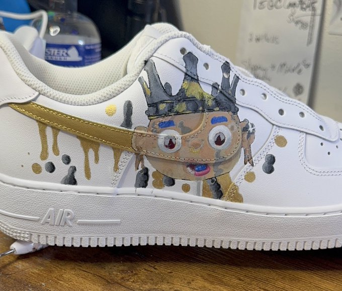 A customized white Nike Air sneaker hand-painted with a quirky cartoon character featuring a crown, dripping gold details, and expressive eyes.