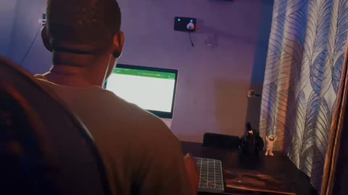 The image shows a man sitting at a desk with a laptop open, likely working or browsing the internet. The background features a window with curtains that allows natural light into the room. There are also two small objects placed near the window - a cup and a vase. The image is related to the post about FHE technology and privacy concerns on the Web3 agents market.