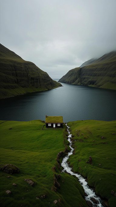 A solitary turf-roofed house sits on lush green slopes beside a winding stream, with a serene fjord flanked by mist-shrouded mountains stretching into the distance—evoking the isolated, family-heritage feel of "Gm Familia."