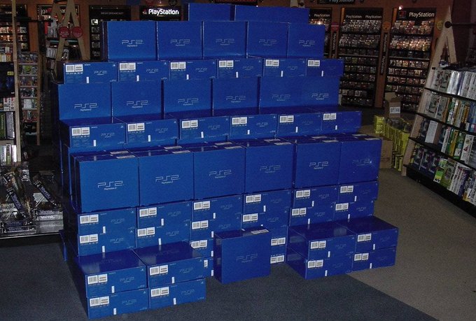 A retail store display featuring a large pyramid-like stack of blue PSP (PlayStation Portable) boxes, with PlayStation branding visible on shelves in the background.